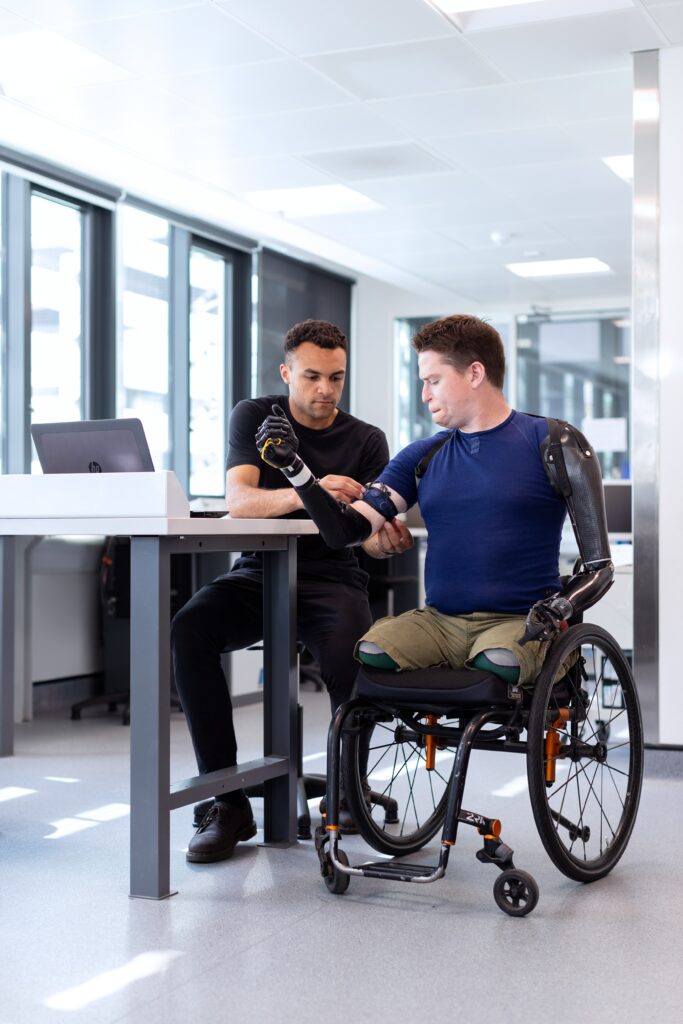 The image by ThisIsEngineering RAEng on Unsplash shows two men working together. One of the men is in a wheelchair with no legs and two prosthetic limbs for arms. The other man who is with him, is a mechanical engineer and sitting beside him in a chair. The engineer is helping with the configuration of the prosthetic limb on his right arm.