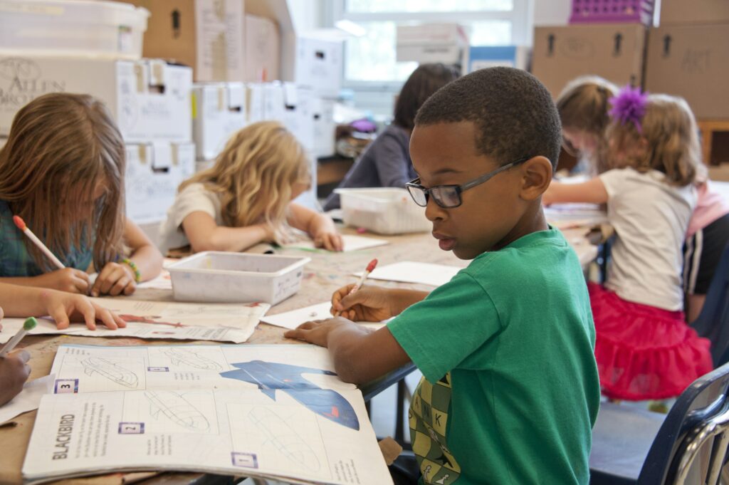 A young boy, roughly age 7, sketching a Blackbird jet in an art class. He is surrounded by his peers doing other various drawings. 