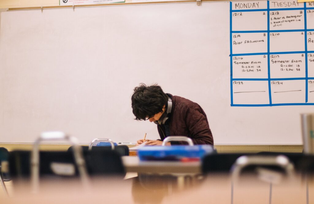 A photo of a male high school student, sitting alone in a classroom with his head down and a pencil in his hand. He seems to be hard at work and focused on his task.