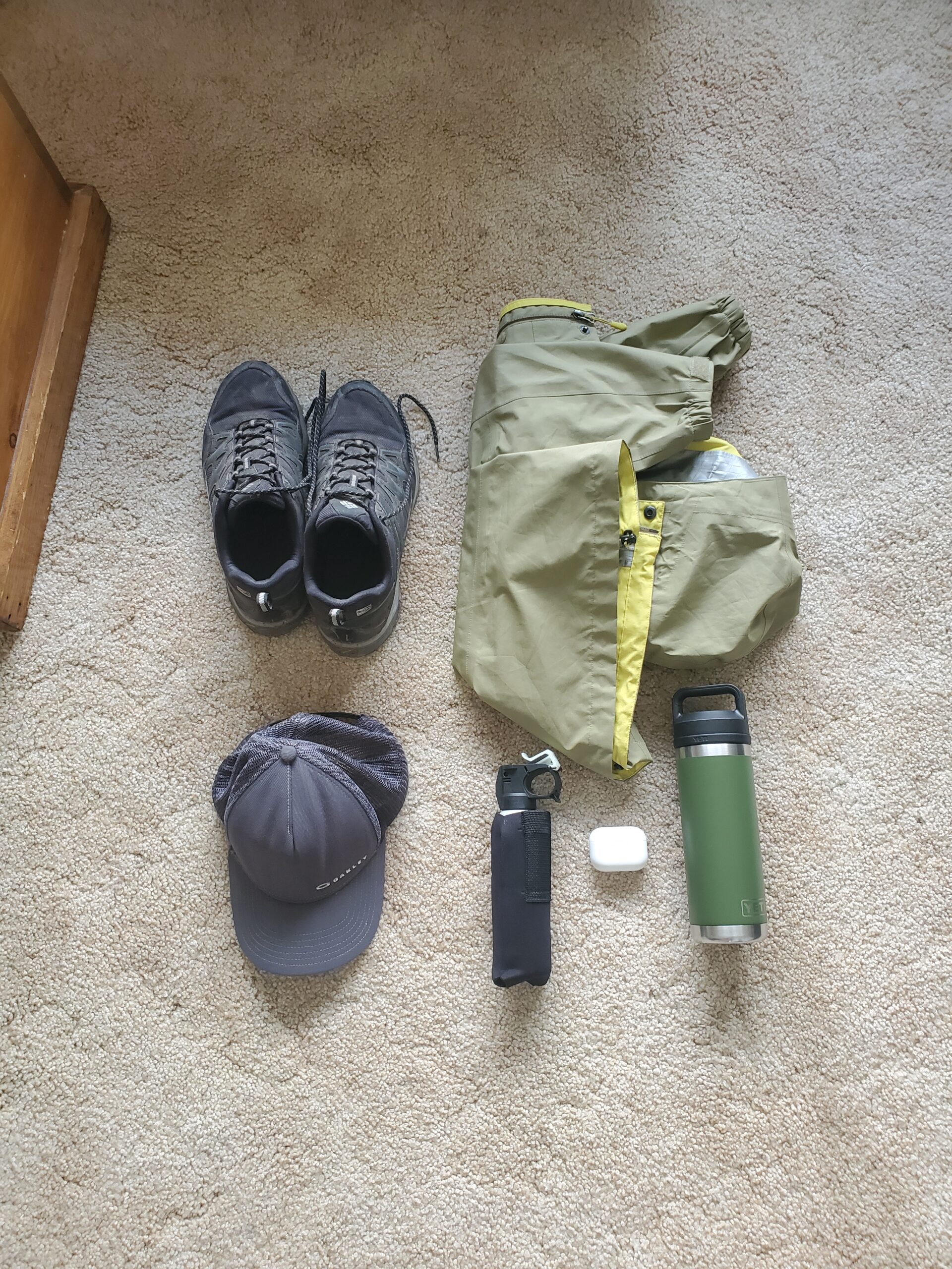 A photo of some basic gear for a short hike. The photo features hiking boots, a raincoat, a hat, bear spray, earbuds and a water bottle.